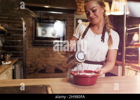 une jeune femme chef debout au comptoir fouette des ingrédients frais dans un bol en plastique avec un batteur électrique. Banque D'Images