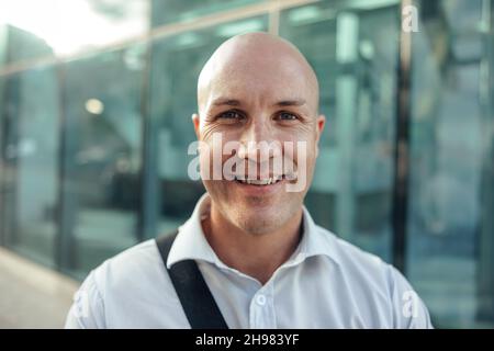 Un homme d'affaires heureux souriant à la caméra tout en se tenant devant un immeuble de bureaux de grande hauteur dans la ville.Entrée adulte moyenne réussie Banque D'Images