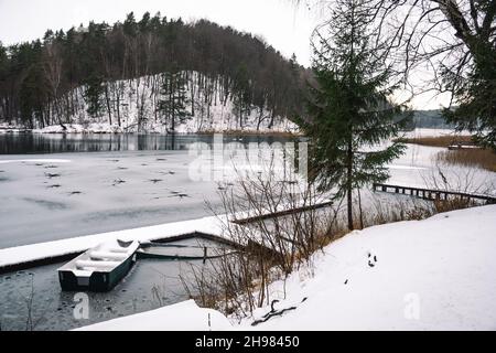 Lac gelé en hiver avec trous, avec jetée, bateaux en bois et sapins couverts de neige, famille de cygnes et forêt en arrière-plan Banque D'Images