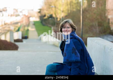 30 yo femme avec un manteau bleu, des lunettes de soleil et un foulard posant au soleil d'hiver dans un parc Banque D'Images