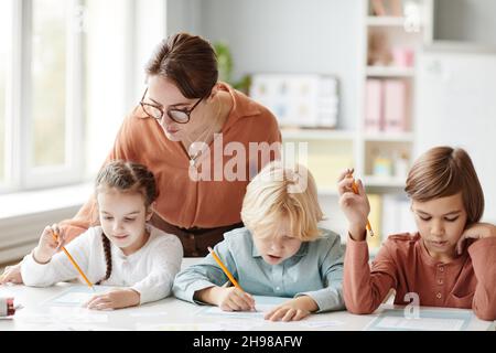 Jeune professeur de lunettes aidant les enfants à faire leurs devoirs à la table dans la salle de classe Banque D'Images