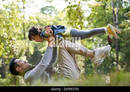 père asiatique allongé sur le dos sur l'herbe ayant un bon moment de soulever la fille dans les airs à l'extérieur dans le parc de la ville Banque D'Images