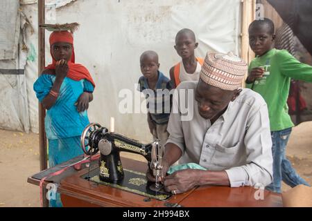 Un tailleur local travaillant sur une machine à coudre ancienne dans une boutique improvisée dans la rue avec des gens locaux et des enfants curieux assis autour Banque D'Images