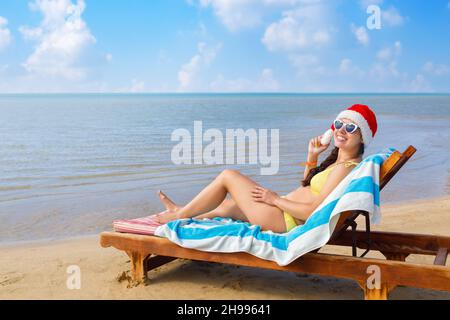 jeune femme gaie en chapeau de père noël et lunettes de soleil couchant sur un transat Banque D'Images