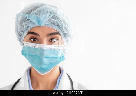 Portrait de femme indienne millénaire médecin en manteau, masque de protection et chapeau, regardant l'appareil photo Banque D'Images