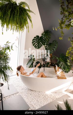 Bonne jeune femme utilisant loofah éponge avec gel douche appliqué tout en se baignant dans la salle de bains moderne décorée avec des plantes tropicales Banque D'Images