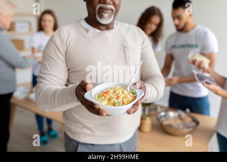 Homme afro-américain senior tenant une assiette avec repas, recevant de la nourriture dans le bureau d'organisation de charité, cultivé, fermé Banque D'Images