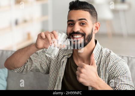 Restez hydraté.Un homme arabe souriant boit de l'eau minérale du verre et montre un geste de pouce à la maison Banque D'Images