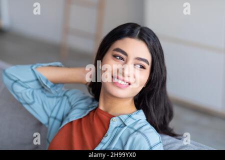 Une femme arabe heureuse s'est penchée sur un canapé, se reposant sur un canapé confortable, tenant une main derrière sa tête, appréciant une journée de détente Banque D'Images