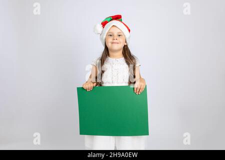 Portrait d'une fille caucasienne souriante en robe blanche et chapeau de gnome de Noël tenant une bannière verte vide isolée sur un fond blanc Banque D'Images