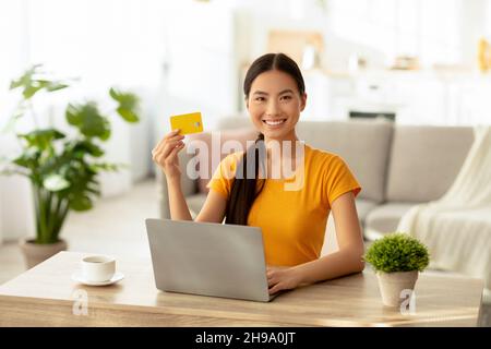 Femme asiatique joyeuse avec carte de crédit à la maison, en commandant de la nourriture ou des vêtements en ligne, assis au bureau Banque D'Images