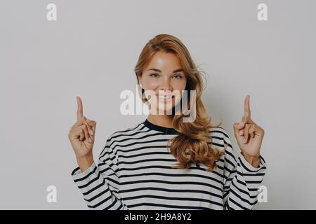 Jeune femme aux cheveux blonds teints souriant agréablement en pointant les index vers le haut Banque D'Images