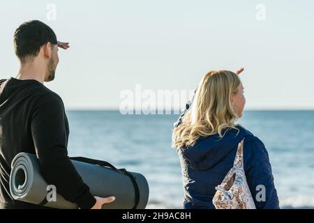 Homme et femme face à la mer tout en portant des tapis de yoga Banque D'Images