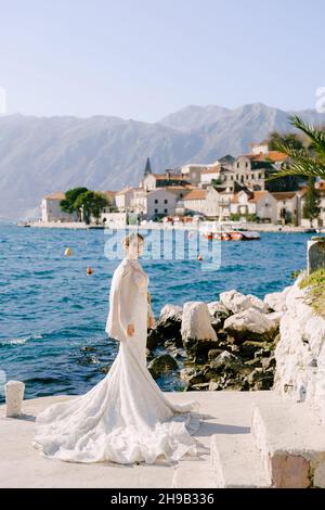 Mariée dans une longue robe blanche de sirène se tient sur un quai au large de la côte de Perast Banque D'Images