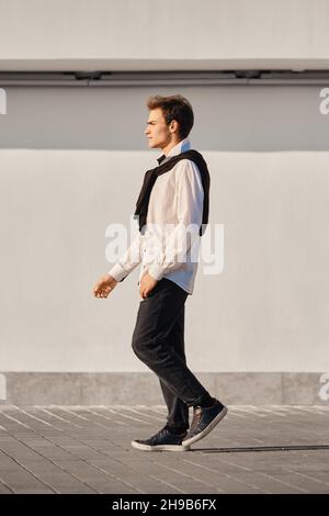 Portrait complet d'un jeune homme en chemise blanche et chandail drappé sur les épaules marche dans la rue Banque D'Images