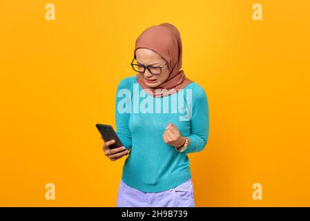 Une jeune femme asiatique en colère regarde l'écran du smartphone isolé sur fond jaune Banque D'Images