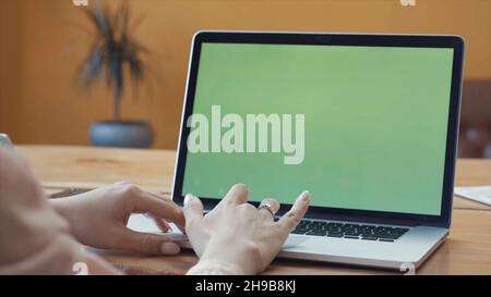 Jeune femme en chemise rose assise à la grande table en bois et dactylographiant sur son ordinateur portable avec la touche chroma écran vert.Écran Chroma Key pour le positionnement de Banque D'Images