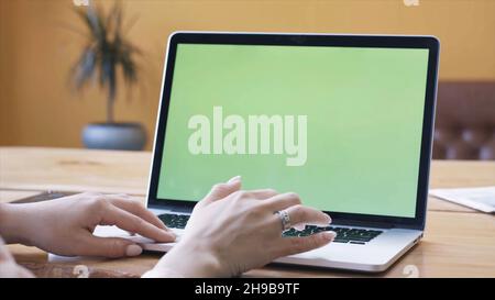 Jeune femme en chemise rose assise à la grande table en bois et dactylographiant sur son ordinateur portable avec la touche chroma écran vert.Écran Chroma Key pour le positionnement de Banque D'Images