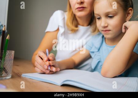 Gros plan de petite fille qui fait ses devoirs avec une jeune mère assise à la table près de la fenêtre. Banque D'Images