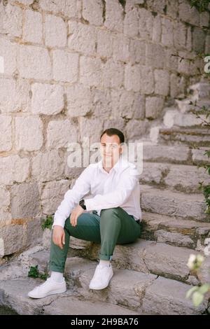 Un homme dans une chemise blanche est assis sur des marches de pierre près d'un mur de briques grises Banque D'Images