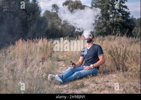 Beau homme de vape appréciant un dispositif électronique de fumée sur l'herbe. Non-fumeurs. Banque D'Images