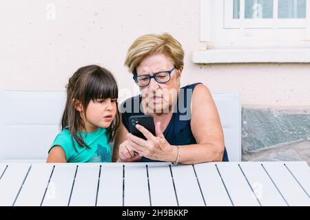 Grand-mère et petite-fille avec un visage étrange consultant le mobile sur une terrasse.Famille, petits-enfants, grands-parents et concept de technologie. Banque D'Images