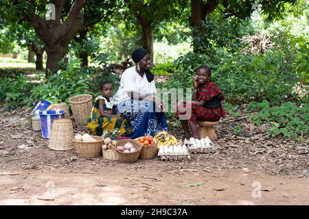 Un grand épicier noir africain souriant, assis avec ses deux filles à côté de la route, attend des clients Banque D'Images