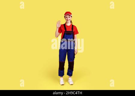 Portrait complet d'une femme sympathique souriante se tenant et se portant la main, en disant bonjour ou au revoir, en portant une combinaison et une casquette rouge.Studio d'intérieur isolé sur fond jaune. Banque D'Images