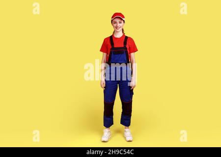 Portrait complet de la femme travailleuse debout et regardant la caméra avec une expression faciale positive, portant une combinaison et une casquette rouge.Studio d'intérieur isolé sur fond jaune. Banque D'Images