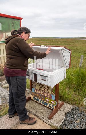 Femme achetant de la nourriture sur un stand de bord de route avec une boîte d'honnêteté à Baltasound sur l'île d'Unst, Shetland. Banque D'Images