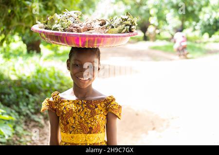 Jolie petite fille africaine noire équilibrant une assiette d'herbes sur sa tête Banque D'Images