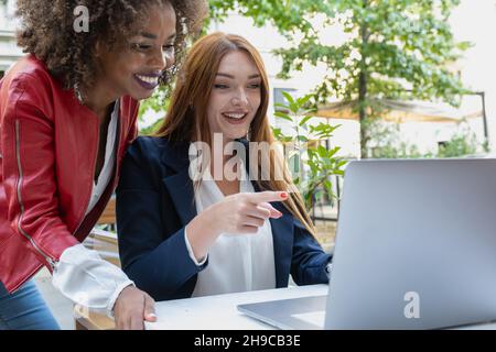 Deux femmes d'affaires analysent des documents sur un ordinateur portable tout en étant assis sur une table dans un lieu de travail extérieur.Femmes cadres au travail montrant un certain succès à Banque D'Images