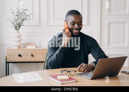 Homme d'affaires afro-américain ayant une agréable conversation téléphonique tout en travaillant sur un ordinateur portable Banque D'Images