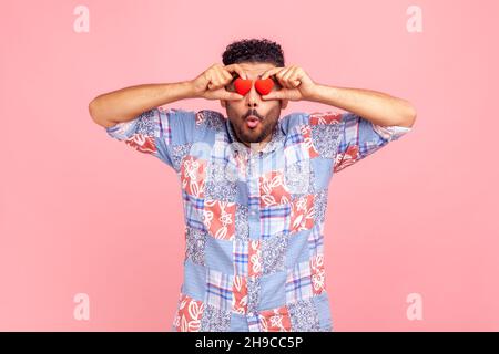 Portrait de l'homme barbu avec une expression faciale étonnante, en gardant les lèvres bouchées, couvrant les yeux avec de petits coeurs rouges, exprimant des émotions choquées.Studio d'intérieur isolé sur fond rose. Banque D'Images