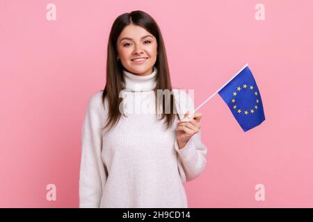 Charmante femme aux cheveux sombres tenant le drapeau syndical européen entre ses mains et regardant sourire à l'appareil photo, portant un pull blanc décontracté.Studio d'intérieur isolé sur fond rose. Banque D'Images