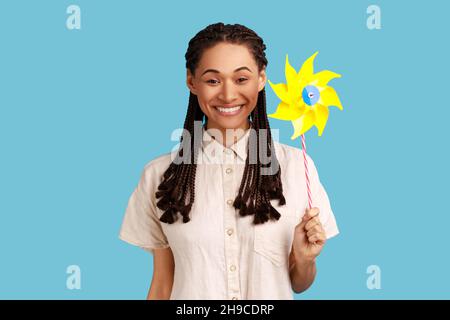 Manuel origami. Portrait d'une femme souriante et insouciante avec des dreadlocks noirs tenant le moulin à vent en papier, le jouet à volant sur bâton, portant une chemise blanche.Studio d'intérieur isolé sur fond bleu. Banque D'Images