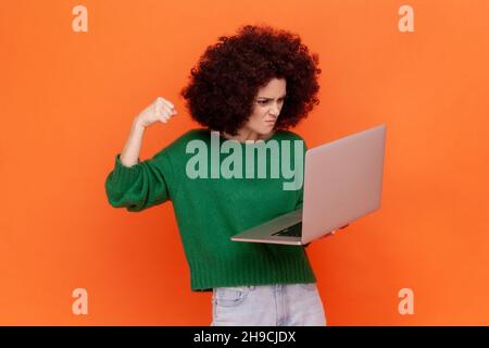 Une femme en colère avec une coiffure afro portant un pull vert décontracté de style travaillant sur un ordinateur portable, a une erreur, veut frapper son poing sur l'écran.Studio d'intérieur isolé sur fond orange. Banque D'Images