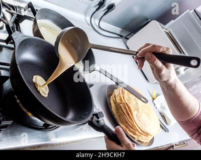 Faire couler une pâte d'un seau en métal sur une poêle graissée rouge-chaude.Le processus de cuisson des crêpes.Un régal traditionnel pour les vacances de Maslenitsa. Banque D'Images