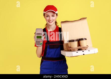 Portrait de la femme de messagerie optimiste tenant le terminal et le café avec boîte à pizza, regardant la caméra avec l'expression heureuse, portant une combinaison et un chapeau.Studio d'intérieur isolé sur fond jaune. Banque D'Images