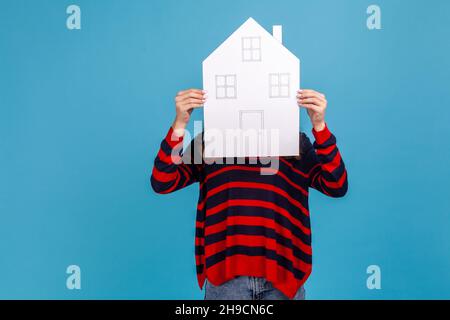 Femme inconnue portant un chandail rayé de style décontracté, cachant son visage derrière la maison de papier avec des fenêtres et des portes dessinées, programme de logement du gouvernement.Studio d'intérieur isolé sur fond bleu. Banque D'Images