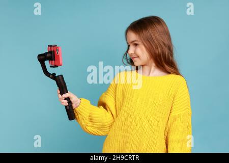 Portrait de petite fille charmante blogueur de diffusion en direct, en utilisant Steadicam et téléphone mobile, portant jaune style décontracté chandail.Studio d'intérieur isolé sur fond bleu. Banque D'Images