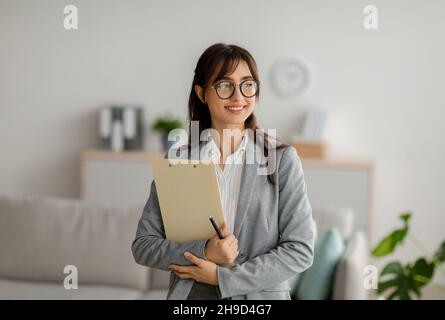Portrait de femme psychothérapeute arabe avec presse-papiers regardant de côté et souriant, debout au bureau moderne, espace libre Banque D'Images
