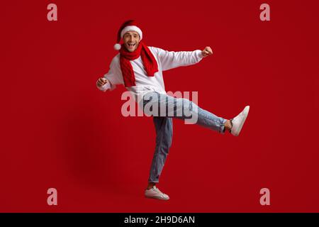 Joyeux jeune homme vêtu d'un chapeau de père Noël et d'une écharpe rouge dansant en studio Banque D'Images