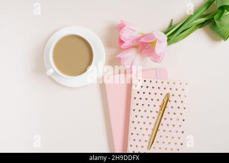 Espace de travail féminin avec fleurs de tulipe roses, journal, tasse de thé et beignet sur fond beige.Pose à plat.Arrière-plan de la vue de dessus. Banque D'Images