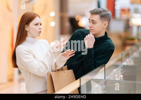 Joyeuse jolie tête rouge jeune femme racontant à l'homme beau histoire d'intérêt debout près de la rampe dans le centre commercial avec intérieur lumineux. Banque D'Images