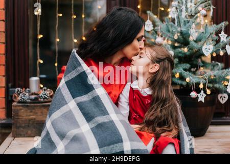 Mère embrassant et enveloppant sa fille souriante dans une couverture sur fond d'arbre de Noël.Ambiance de Noël.Vacances parentales individuelles en solo.Temps familial CLO Banque D'Images