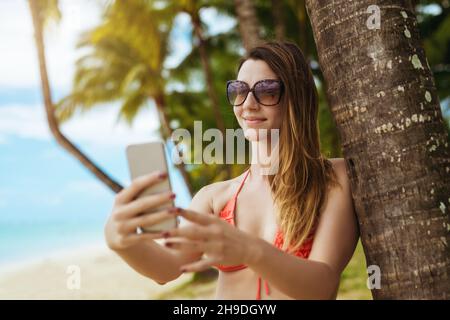 Belle femme en mode bikini prenant selfie sur un magnifique fond tropical.Appel vidéo avec des amis Banque D'Images