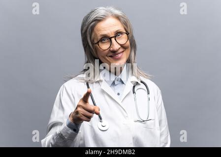 Souriante mature cheveux gris femme médecin en robe médicale blanche, avec stéthoscope isolé sur fond gris points doigt sur vous, vous choisir, MD paie l'attention à votre santé et l'assurance médicale Banque D'Images