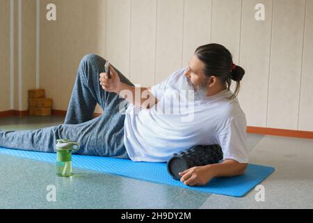 Caucasien barbu, homme long cheveux avec rouleau en mousse et shaker avec l'eau posée sur le tapis de yoga et en utilisant son téléphone portable.Concept de formation en ligne. Banque D'Images