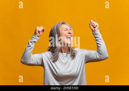 Une femme moderne mûre excitée et surjoyeuse, qui célèbre la réalisation de ses objectifs, élève des poings isolés sur une femme jaune à cheveux gris, se réjouit, criant oui dans l'euphorie, la victoire et la victoire Banque D'Images